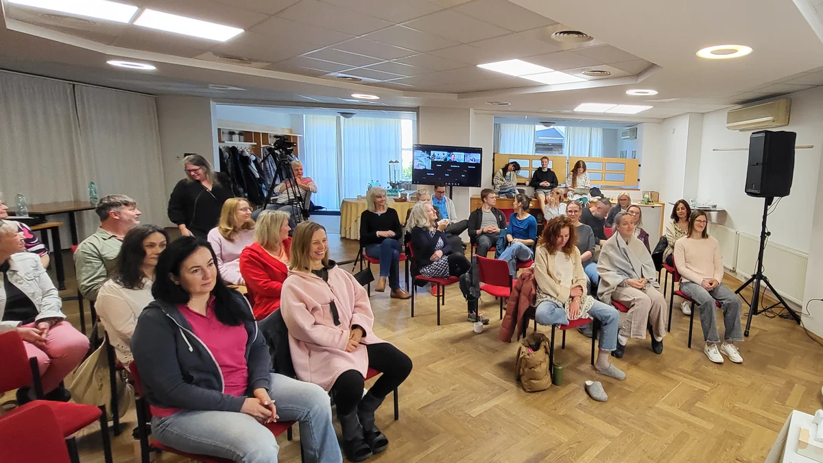 Sál s účastníky v půlkruhu, na obrazovce lidé připojení online